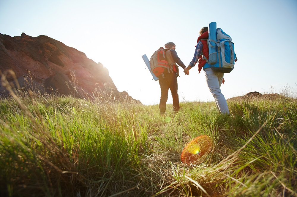 Romantic adventure of young tourist couple, low angle of man and woman going uphill holding hands towards bright sunlight on hiking path in mountains